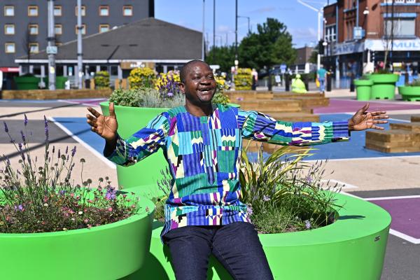 A cheerful man in a colorful shirt sits outdoors near green planters with flowers.